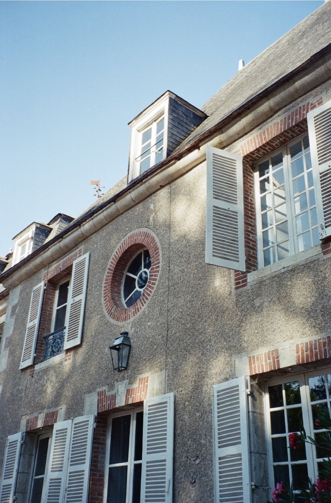 The stone and brick façade from below, with white shutters, a round window and dormers against a blue sky