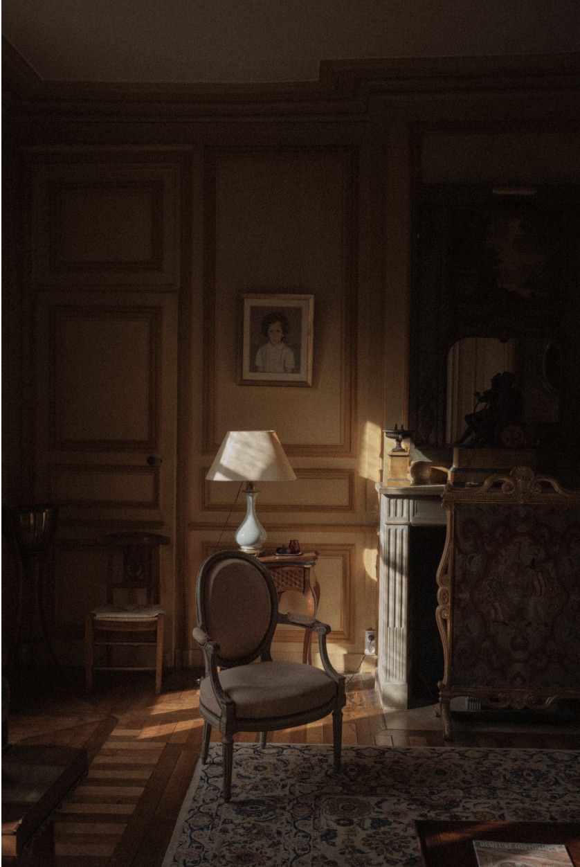 A wood-panelled salon with an armchair, porcelain lamp and marble fireplace in afternoon light