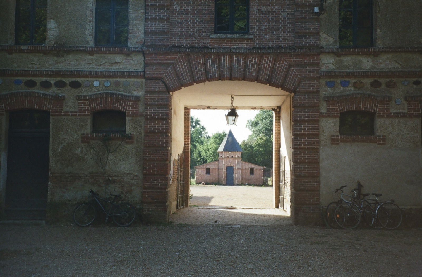 Looking through a brick archway with bicycles parked either side, out to a small turreted building across the gravel courtyard