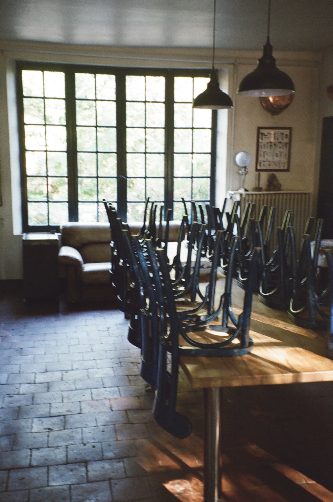 A kitchen with a long wooden table, stacked black chairs, terracotta floor tiles and tall windows